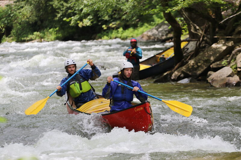 The image shows three people in canoes navigating a whitewater river. Two are in a red canoe in the foreground, paddling through the rapids. The person in the front wears a white helmet and blue jacket, while the person in the back also wears a white helmet and a blue jacket. A third person is further back in a yellow and black canoe. The river is turbulent with white water, and the banks are lined with trees and rocks.